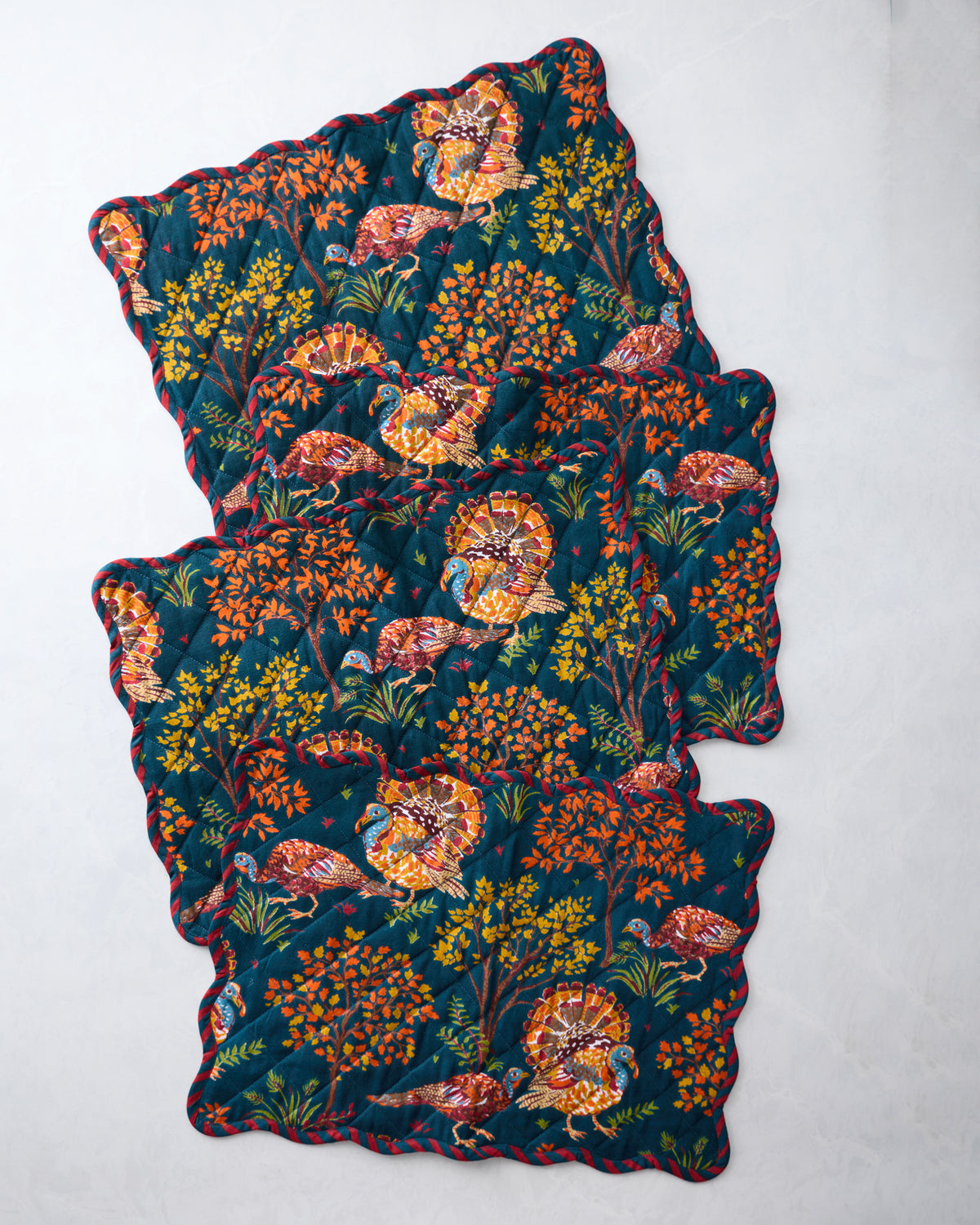 Four navy quilted placemats with scalloped edges, printed with colorful turkeys and foliage, arranged overlapping.
