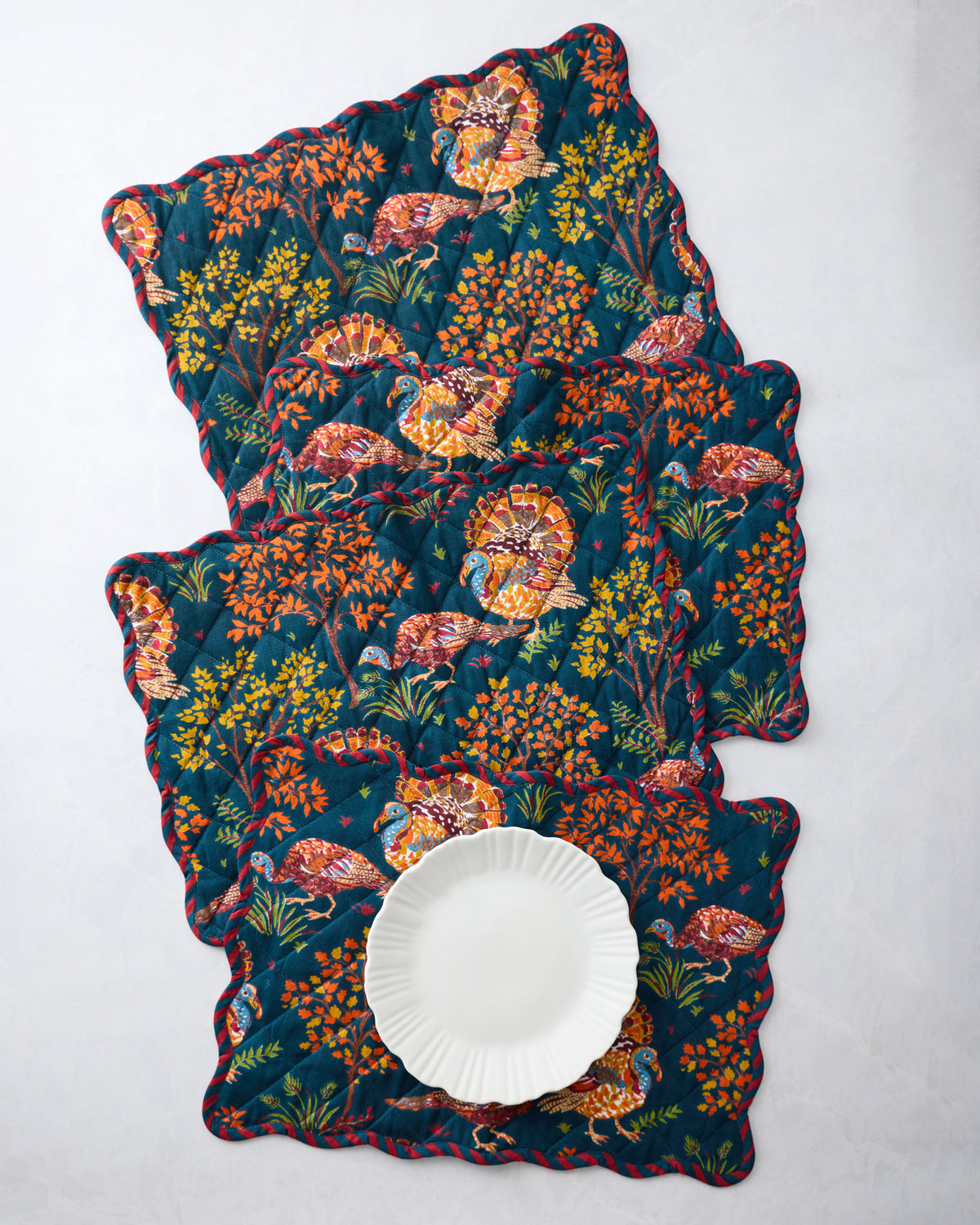 Four navy quilted scalloped placemats with colorful turkey and foliage print, arranged with a white plate on the front placemat.