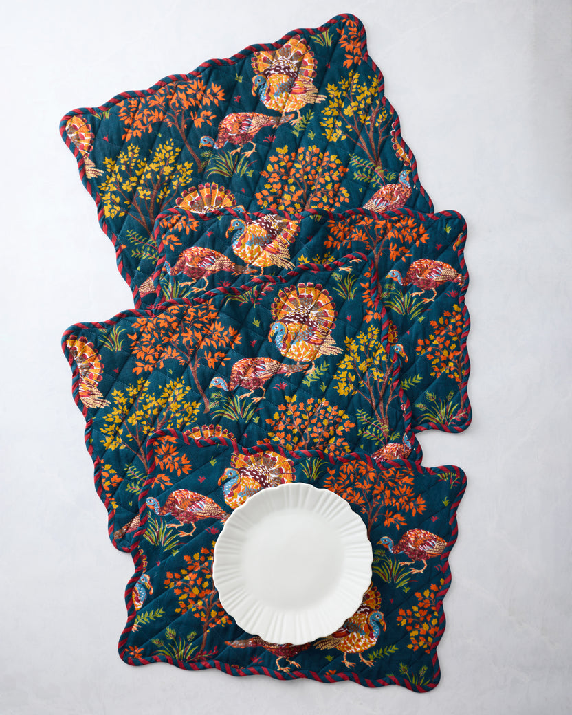 Four navy quilted scalloped placemats with colorful turkey and foliage print, arranged with a white plate on the front placemat.