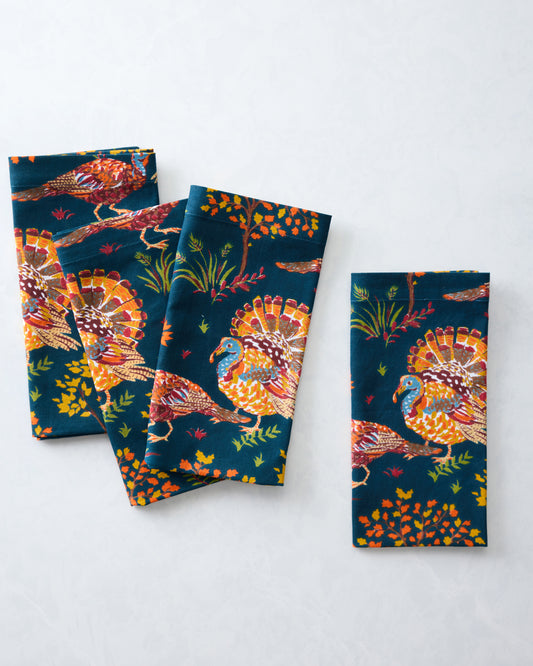 Four navy dinner napkins arranged overlapping, printed with colorful turkeys, trees, leaves, and grasses.
