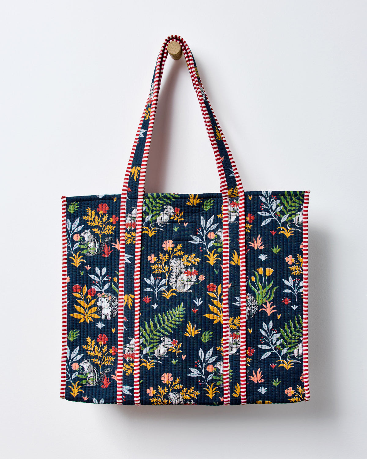 Indigo traveler tote displayed against a light background, decorated with woodland animals, ferns, and leaves and finished with red-and-white striped trim.