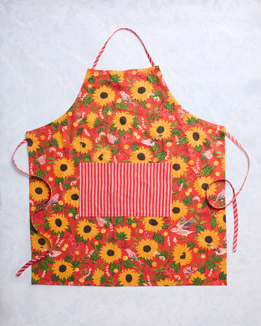 Orange apron laid flat with a sunflower, leaf, and bird print, red-and-white striped pocket, and tie straps.