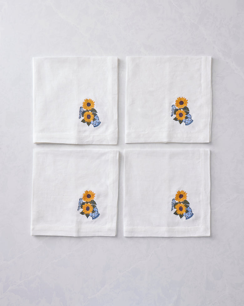 Four white napkins arranged in a square, each embroidered with sunflowers in a blue vase and a bird.