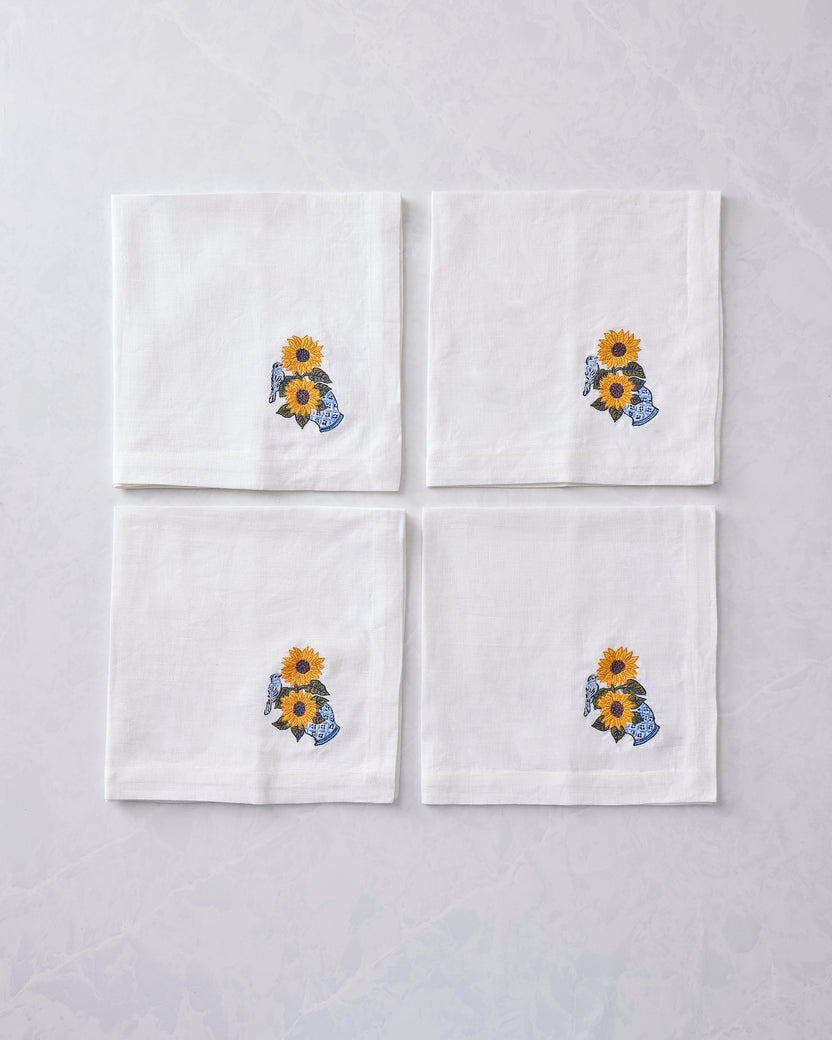 Four white napkins arranged in a square, each embroidered with sunflowers in a blue vase and a bird.