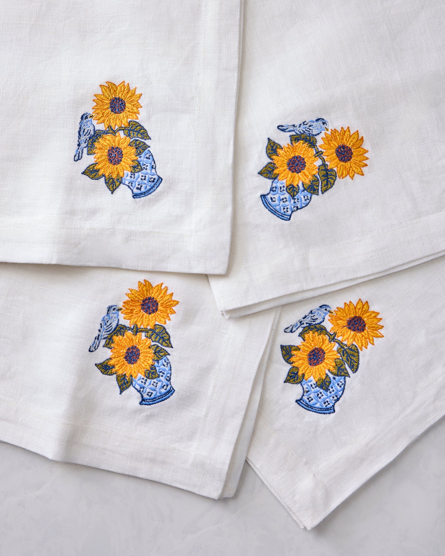 Close-up of four white napkins featuring embroidered sunflowers in blue vases with birds.