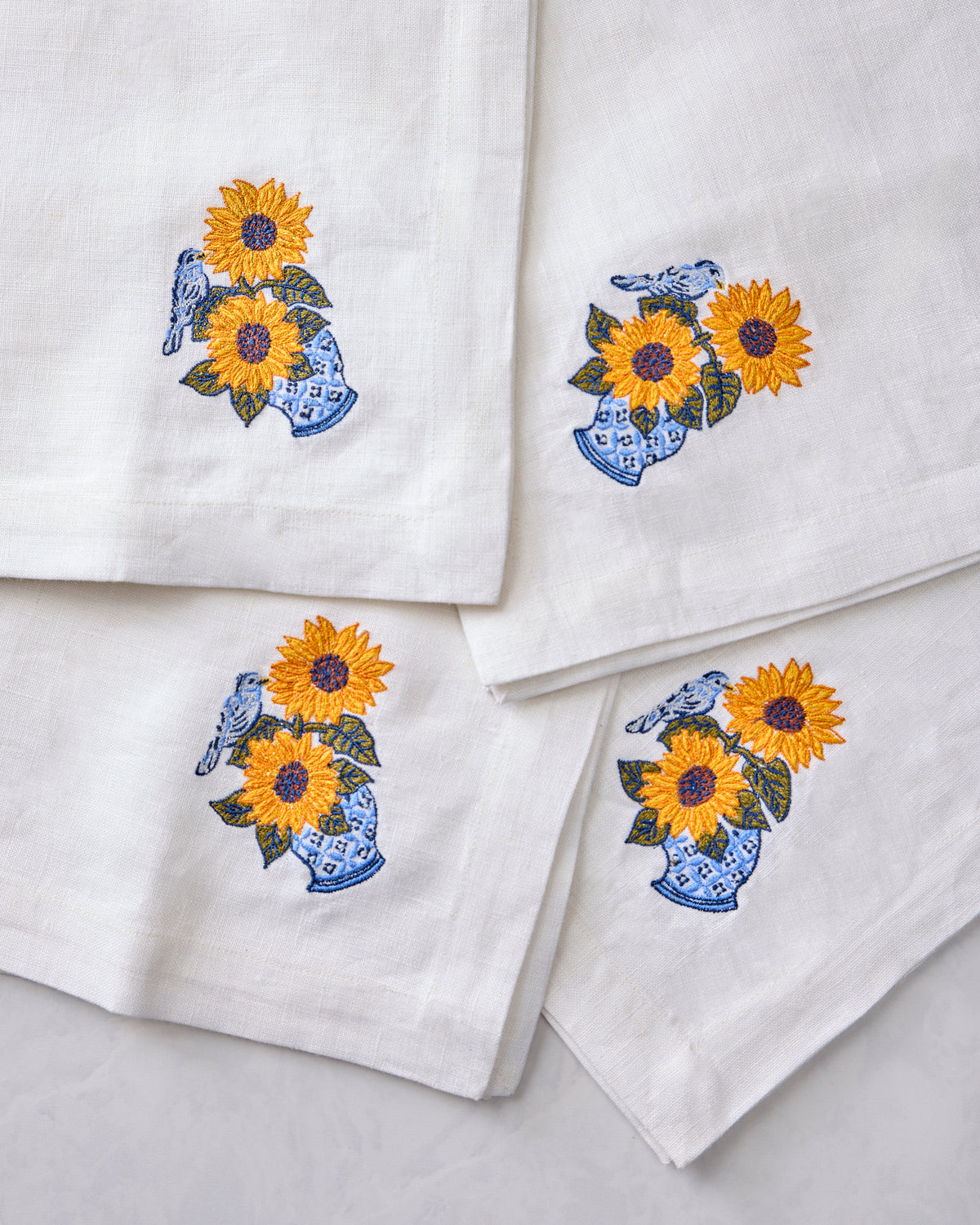 Close-up of four white napkins featuring embroidered sunflowers in blue vases with birds.