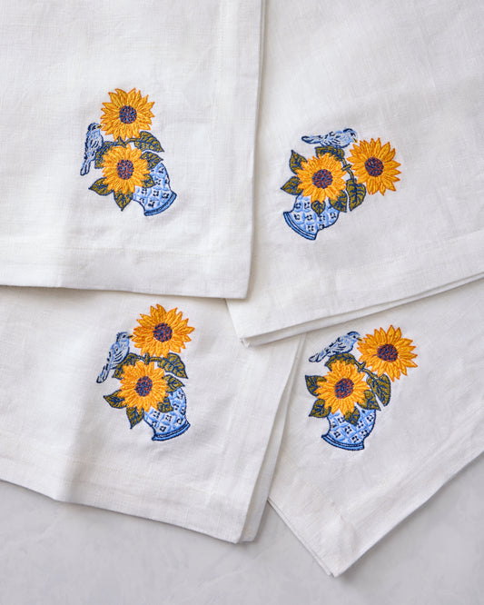 Close-up of four white napkins featuring embroidered sunflowers in blue vases with birds.