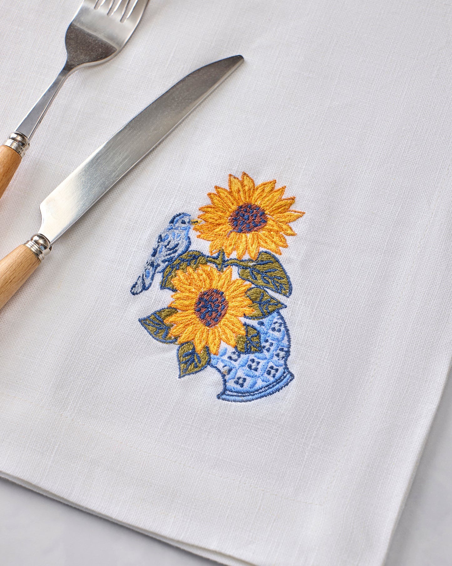Oversized white napkin embroidered with two sunflowers and blue floral leaves, shown with a fork and knife.