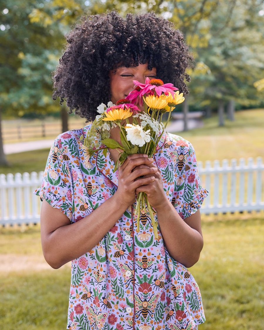 Front view of the lavender Bee Tapestry ruffle short pajama set with flutter sleeves and bee-and-floral print, shown with flowers.