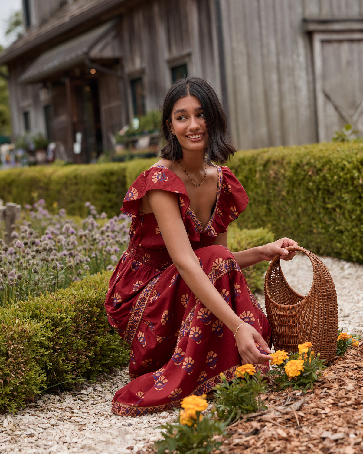 Model wearing the Red Rock Cordelia Dress in a garden, showing its deep V-neck, ruffle sleeves, smocked bodice, patterned maxi skirt, and border trim.