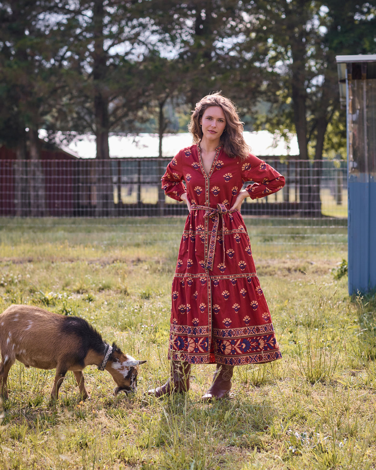 Front view of the Maybelle red patterned maxi dress with long sleeves, waist belt, and decorative trim, shown beside a goat.