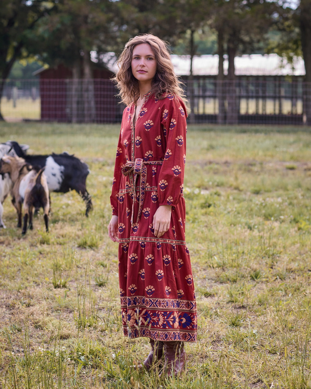 Person wearing the red Canyon Wildflower Maybelle maxi dress with long sleeves and an adjustable waist belt in a grassy outdoor setting.