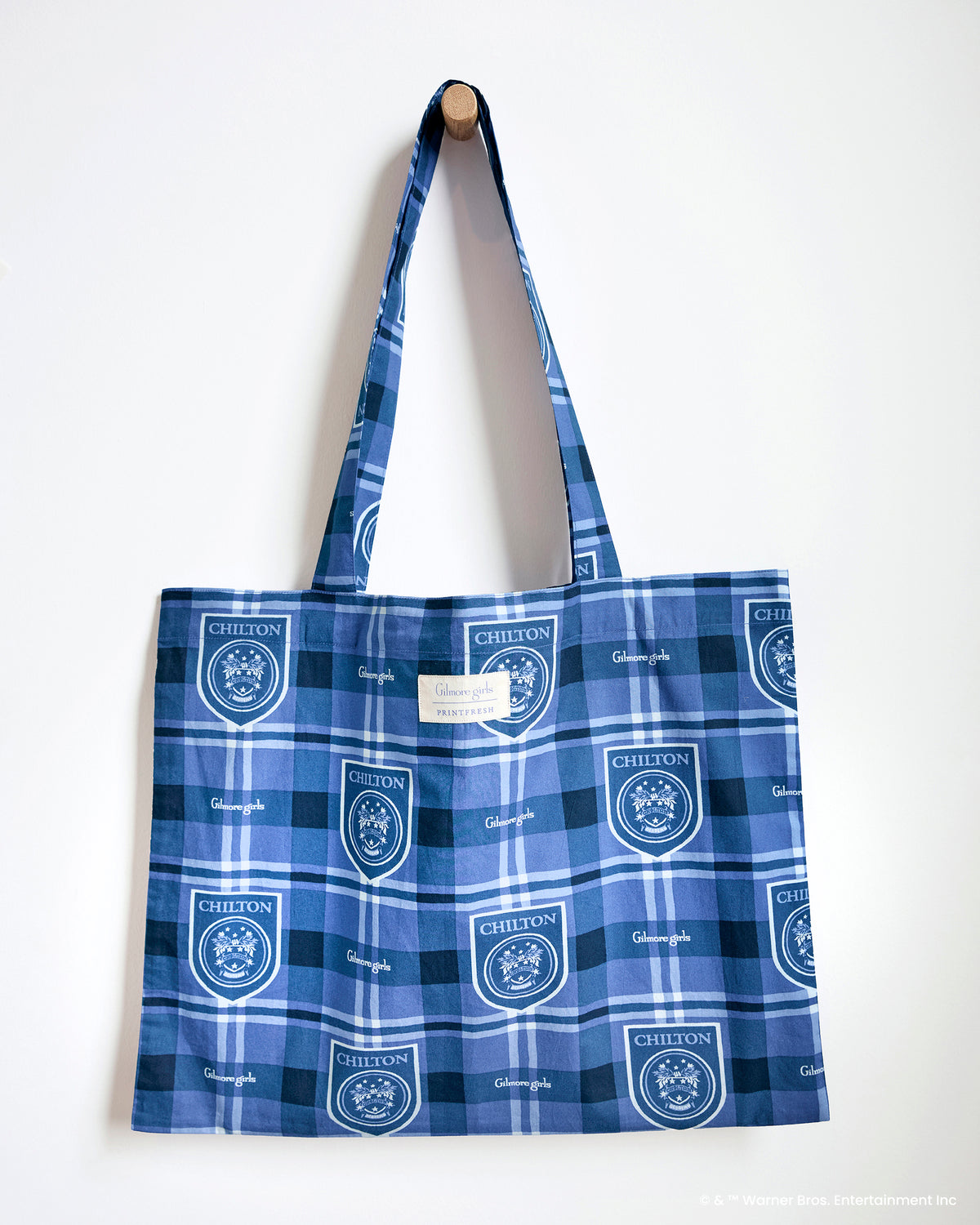 Blue plaid tote bag hanging from a wall peg, with long handles and repeated Chilton crest and Gilmore Girls prints.