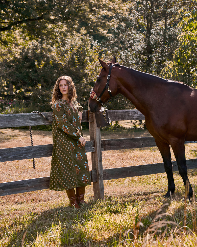 Side view of the deep olive Tallulah dress with floral print, shown beside a horse in a fenced outdoor setting.