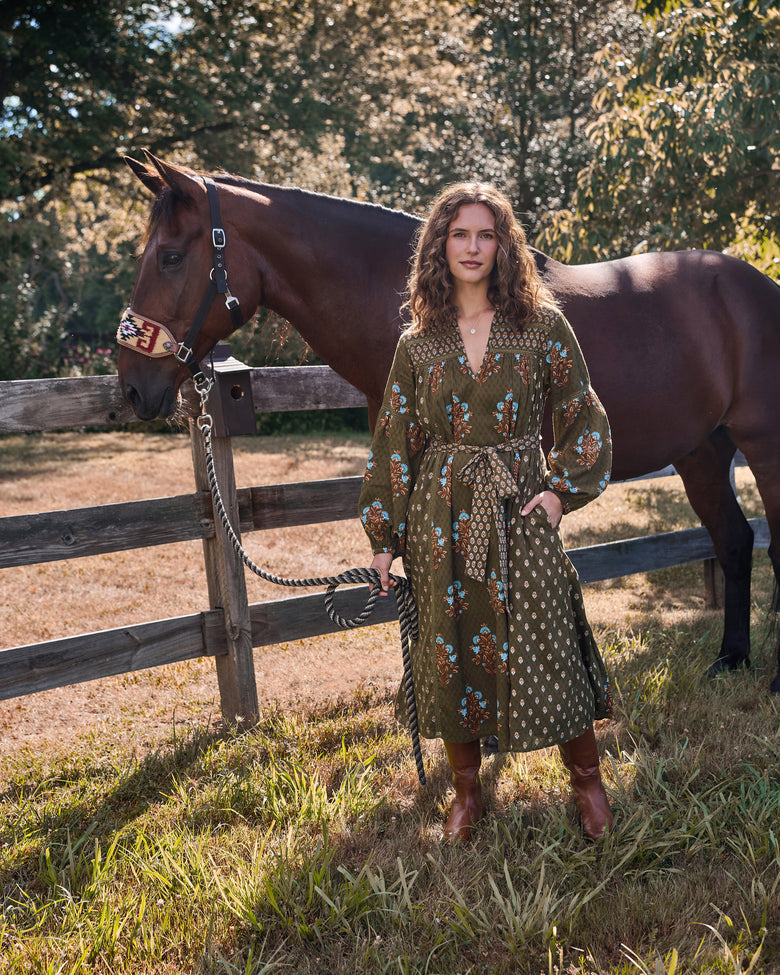 Full view of the deep olive Tallulah dress with floral print and tied waist sash, shown beside a horse.
