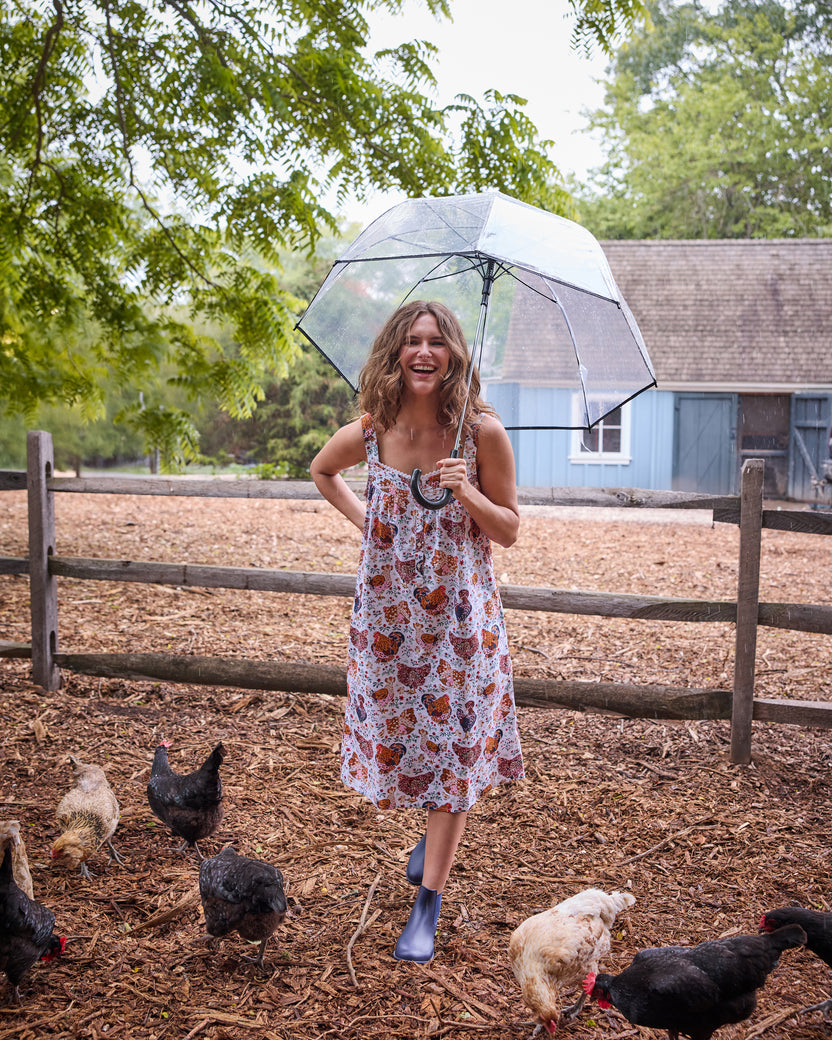 Person wearing a floral chicken-print nightgown and holding a clear umbrella among chickens outdoors.