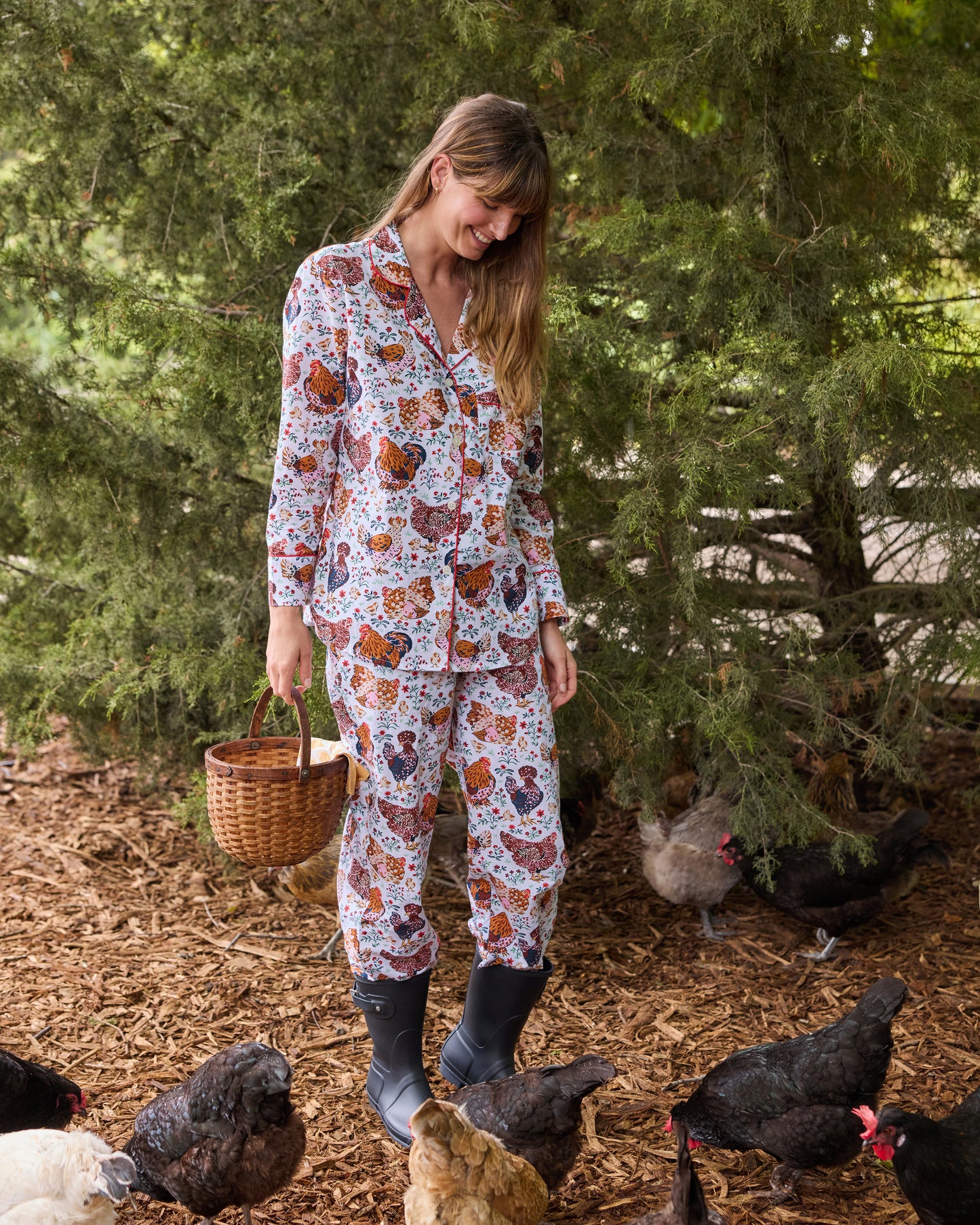 Person wearing a long-sleeved floral chicken-print pajama set with pants and boots among chickens outdoors.