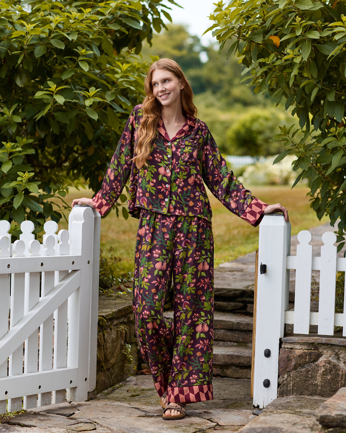 Person wearing the plum Pomme & Poire pajama set with a long-sleeved button-front top and wide-leg pants, standing by a white garden gate; the set has a fruit-and-leaf print with contrasting cuffs and pant hems.