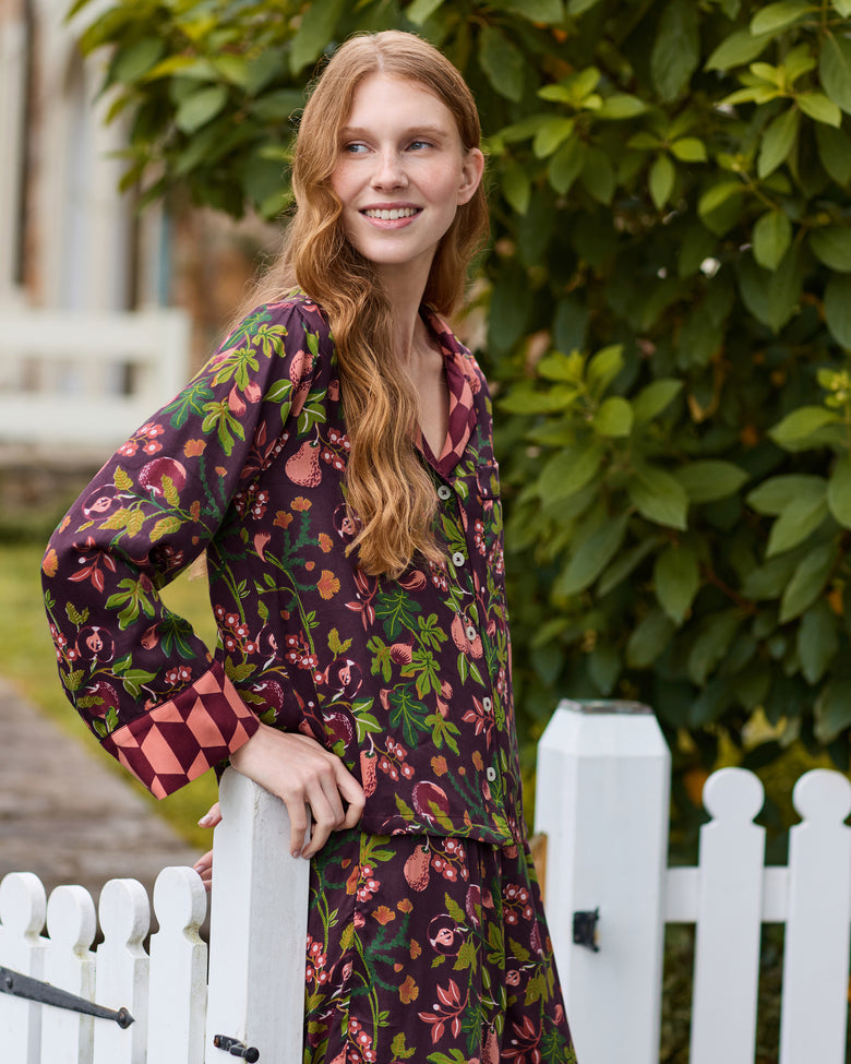 Close lifestyle view of the plum Pomme & Poire pajama top showing its button-front design, collar, chest pocket, fruit-and-leaf print, and contrasting geometric cuffs.
