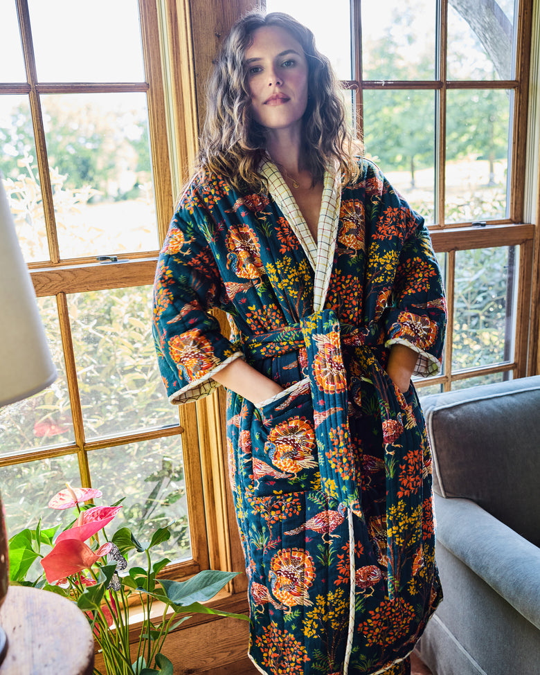 Person wearing a navy Turkey Trot quilted robe with turkey and autumn foliage print; contrasting plaid lining is visible at the collar and cuffs.