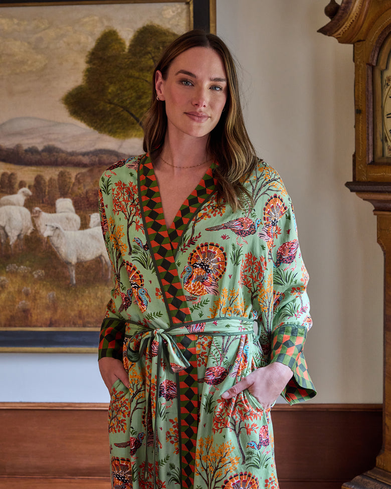 Person wearing a soft sage Turkey Trot long robe with turkey and autumn foliage print, contrasting geometric trim, belt, and pockets.