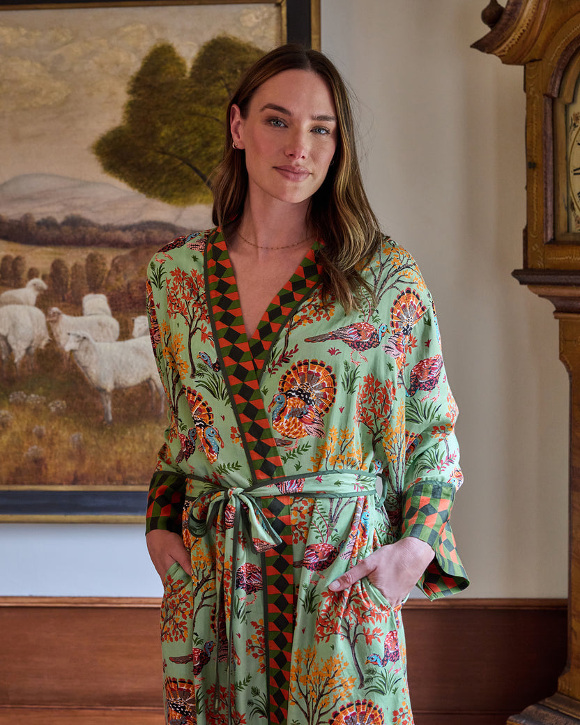 Person wearing a soft sage Turkey Trot long robe with turkey and autumn foliage print, contrasting geometric trim, belt, and pockets.
