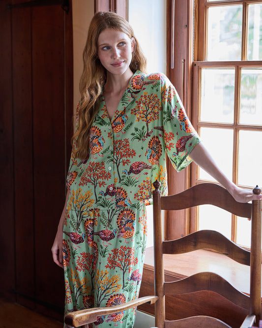 Person wearing a sage button-front pajama set with a colorful turkey and autumn foliage print, standing beside a wooden chair.