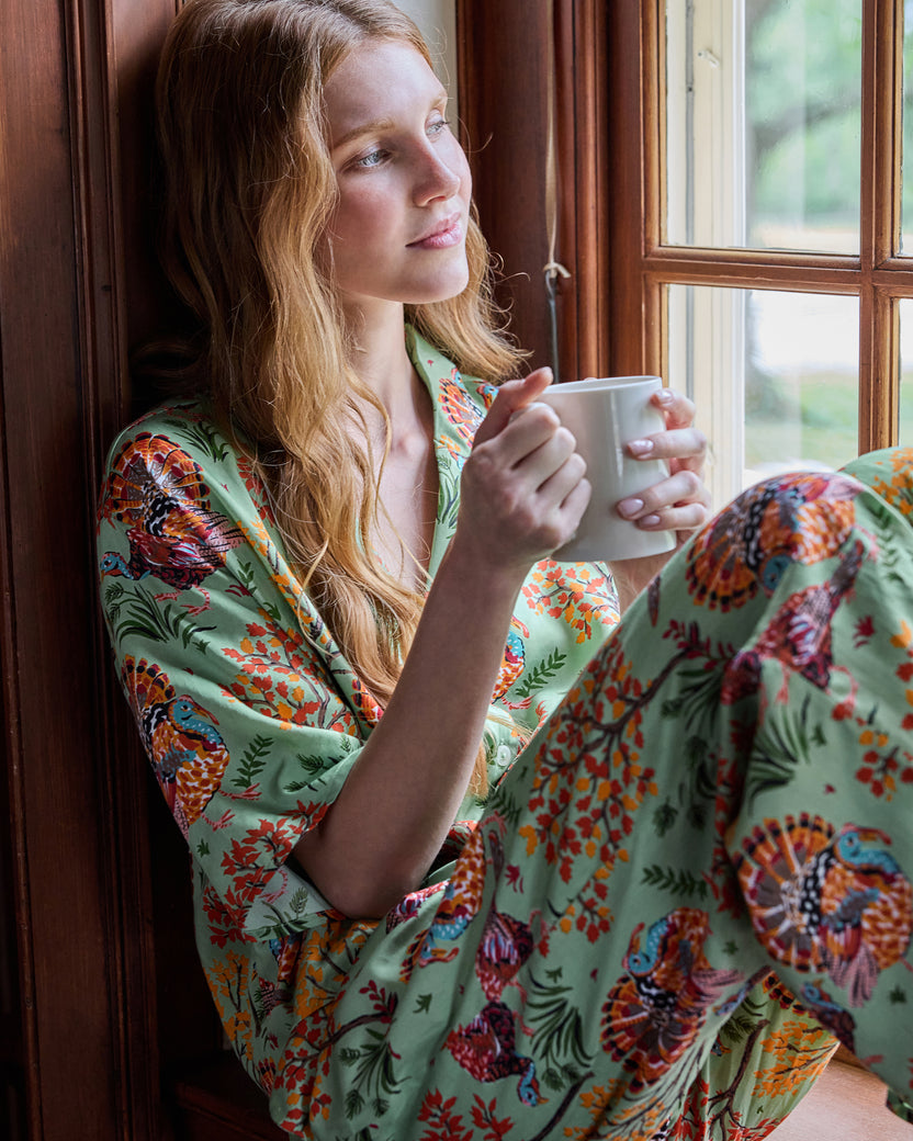 Person wearing a sage turkey-print pajama set and holding a white mug while seated beside a window.