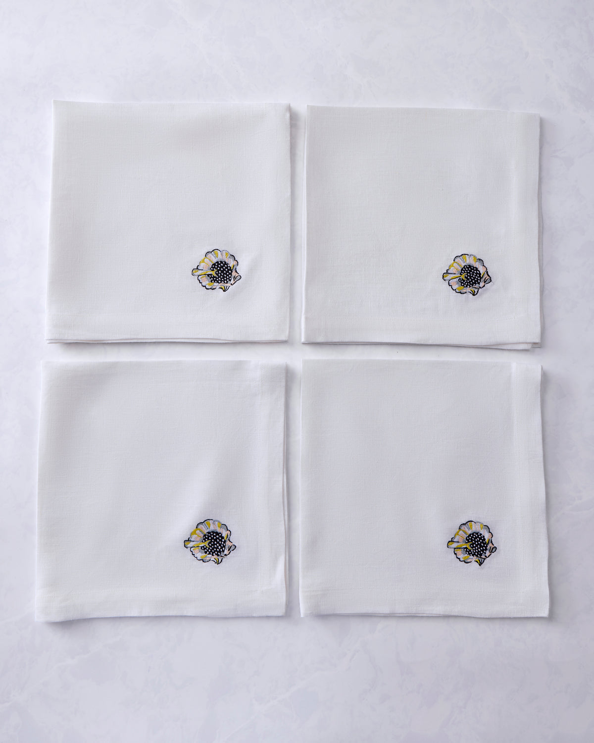 Four white dinner napkins arranged in a square, each embroidered with a shell motif.