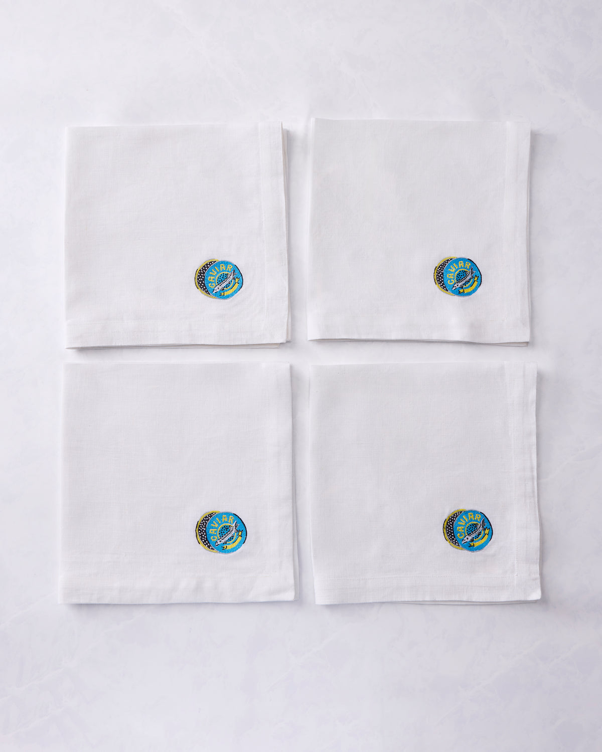 Set of four folded dinner napkins arranged in a square, each with an embroidered caviar design.