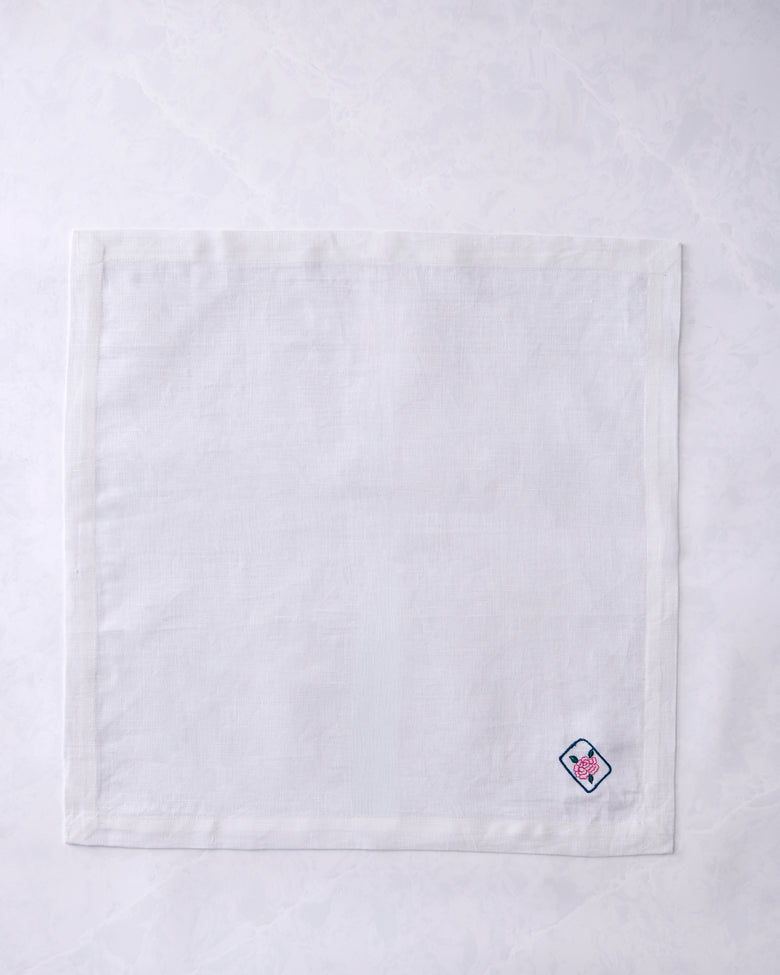 White dinner napkin lying flat with a small embroidered outlined tile containing a pink flower near the lower right corner.