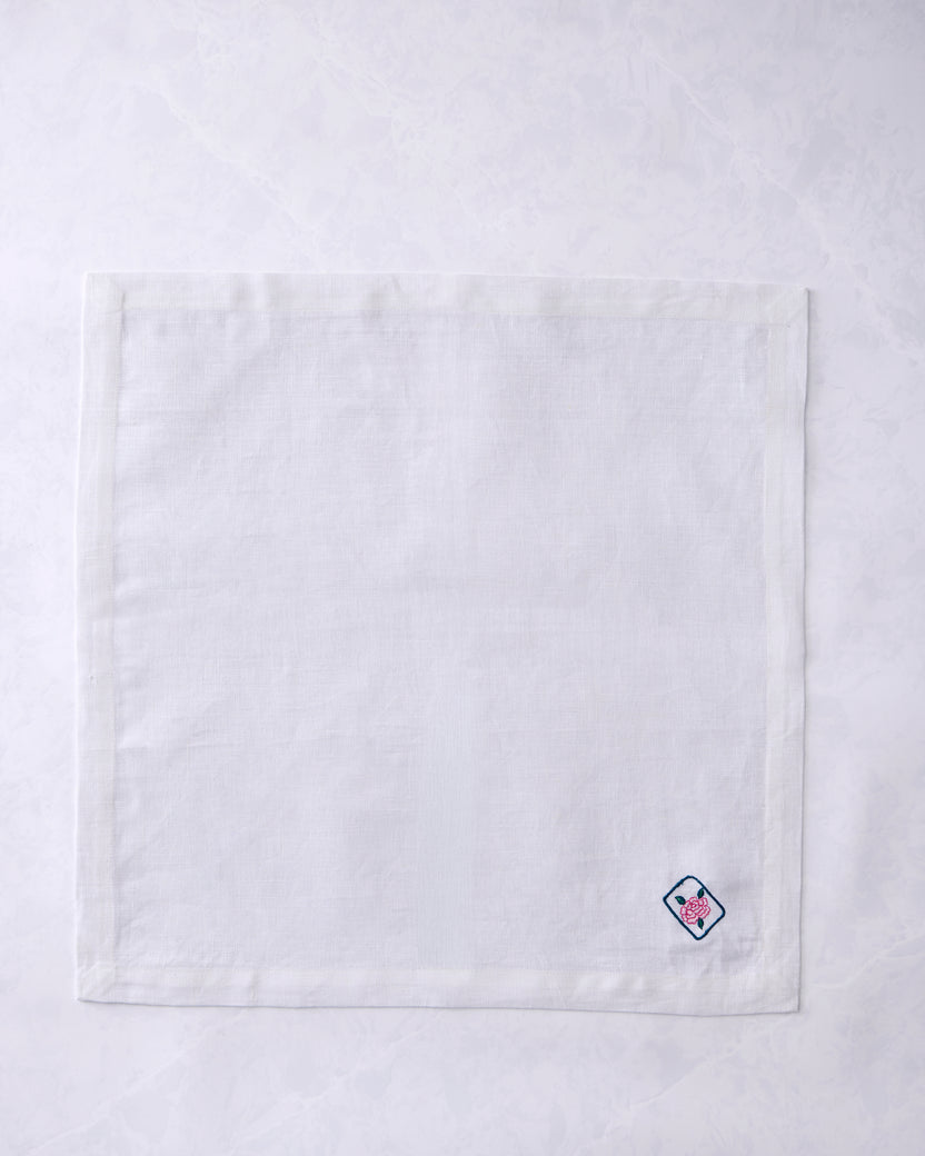 White dinner napkin lying flat with a small embroidered outlined tile containing a pink flower near the lower right corner.