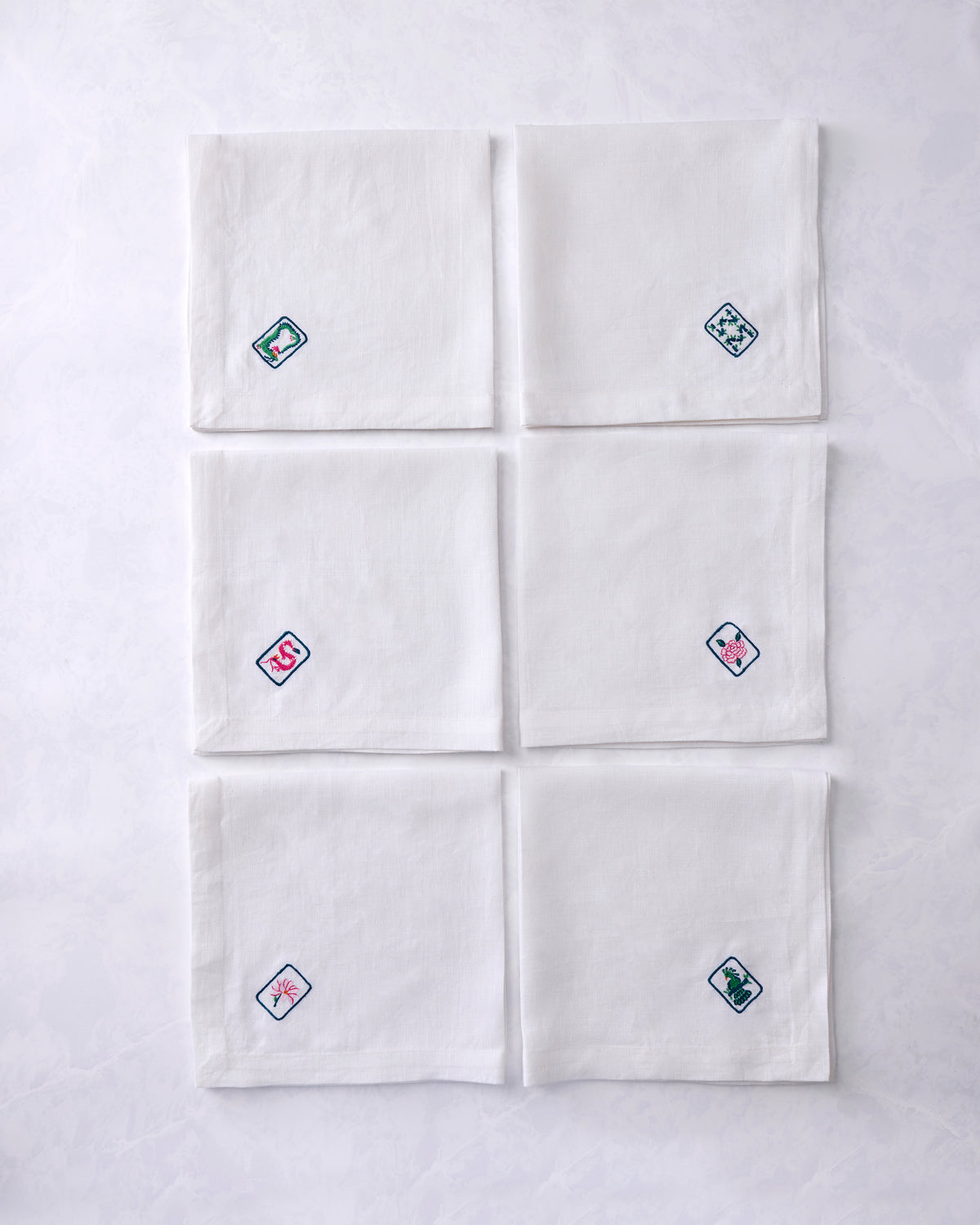 Set of six white dinner napkins arranged in two columns, each embroidered with a different outlined tile motif featuring floral or animal designs.