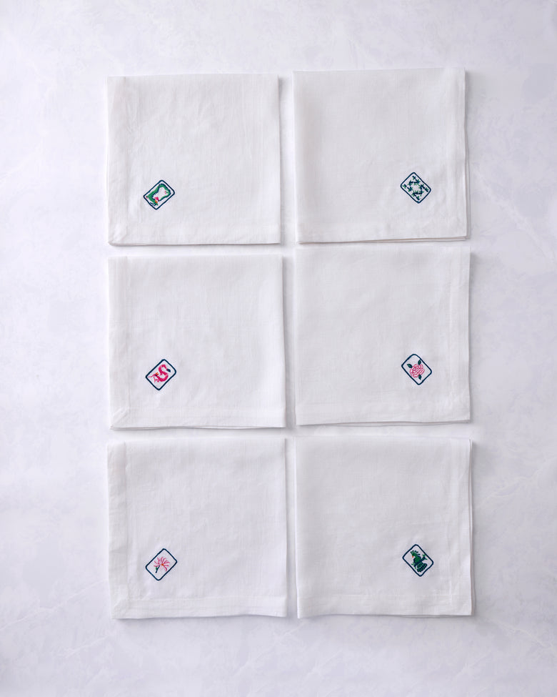 Set of six white dinner napkins arranged in two columns, each embroidered with a different outlined tile motif featuring floral or animal designs.