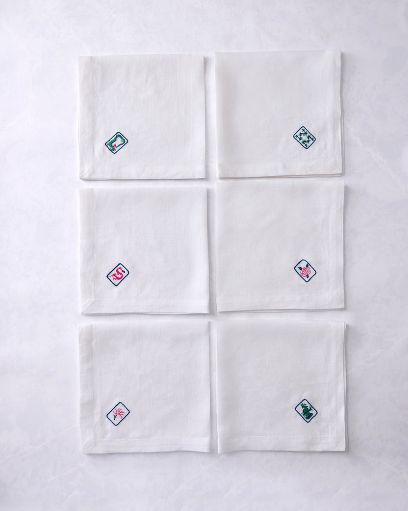 Set of six white dinner napkins arranged in two columns, each embroidered with a different outlined tile motif featuring floral or animal designs.
