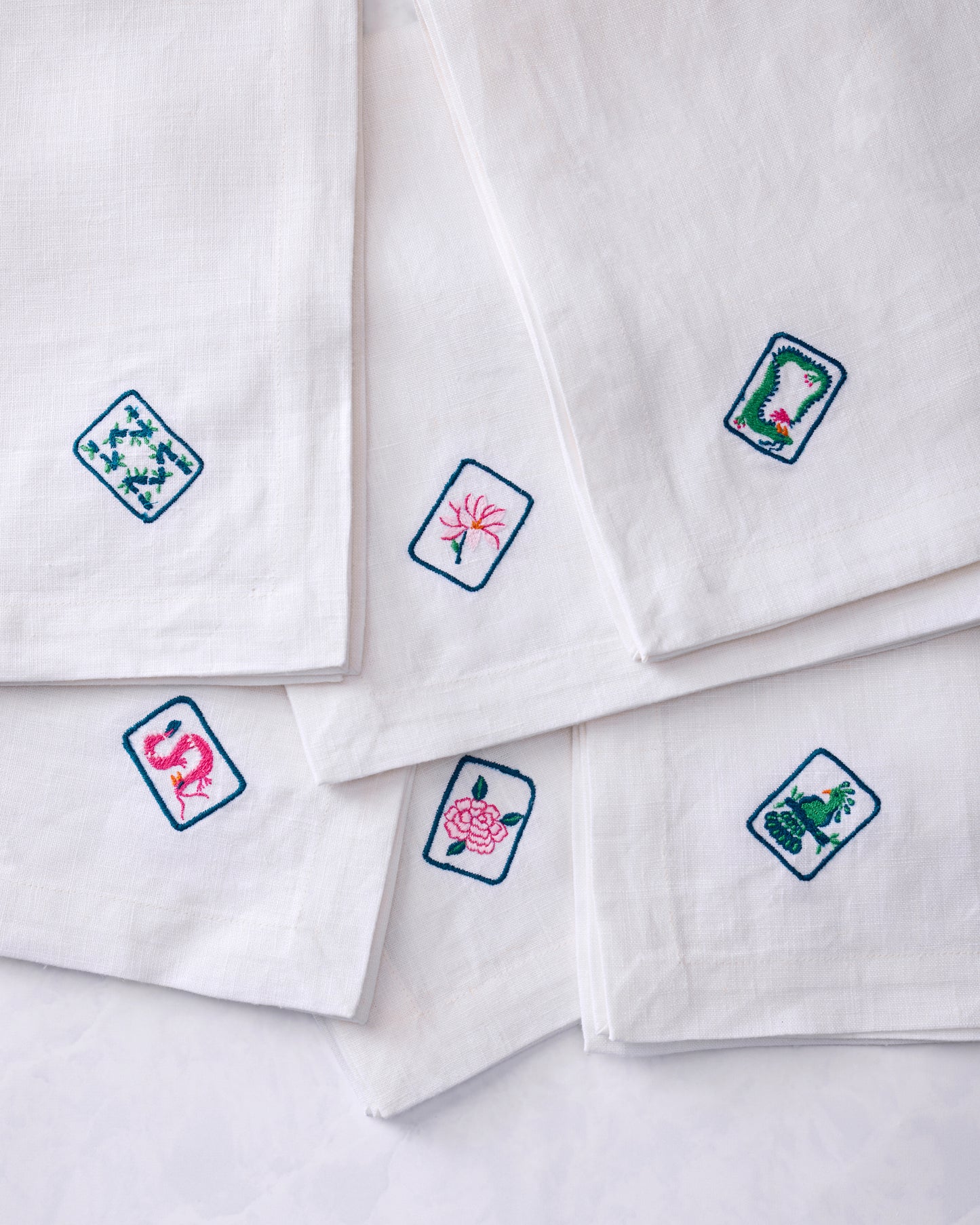 Six white dinner napkins arranged in an overlapping display, each showing a different colorful embroidered tile motif.