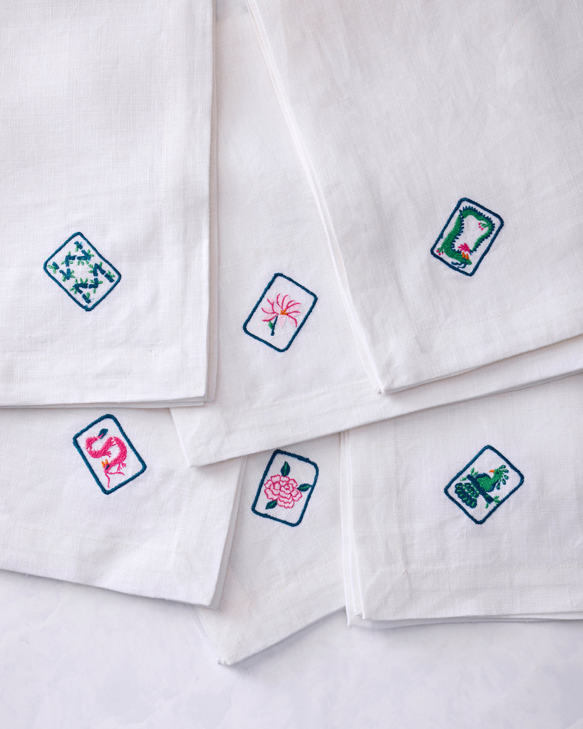 Six white dinner napkins arranged in an overlapping display, each showing a different colorful embroidered tile motif.