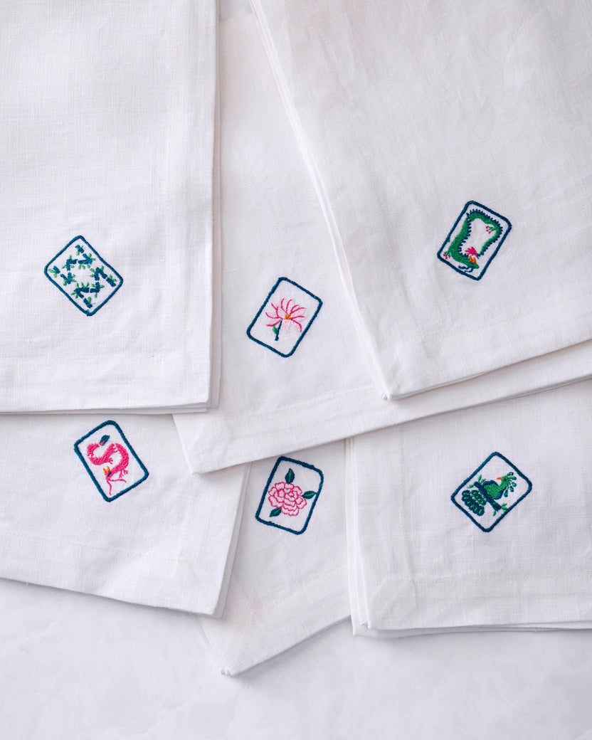 Six white dinner napkins arranged in an overlapping display, each showing a different colorful embroidered tile motif.