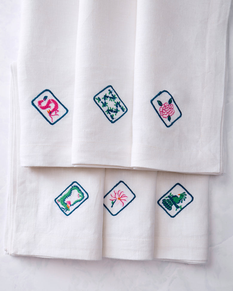 Close-up of six white dinner napkins displaying different colorful embroidered tile motifs, including a pink flamingo, leafy design, pink flower, floral wreath, flower, and green animal motif.