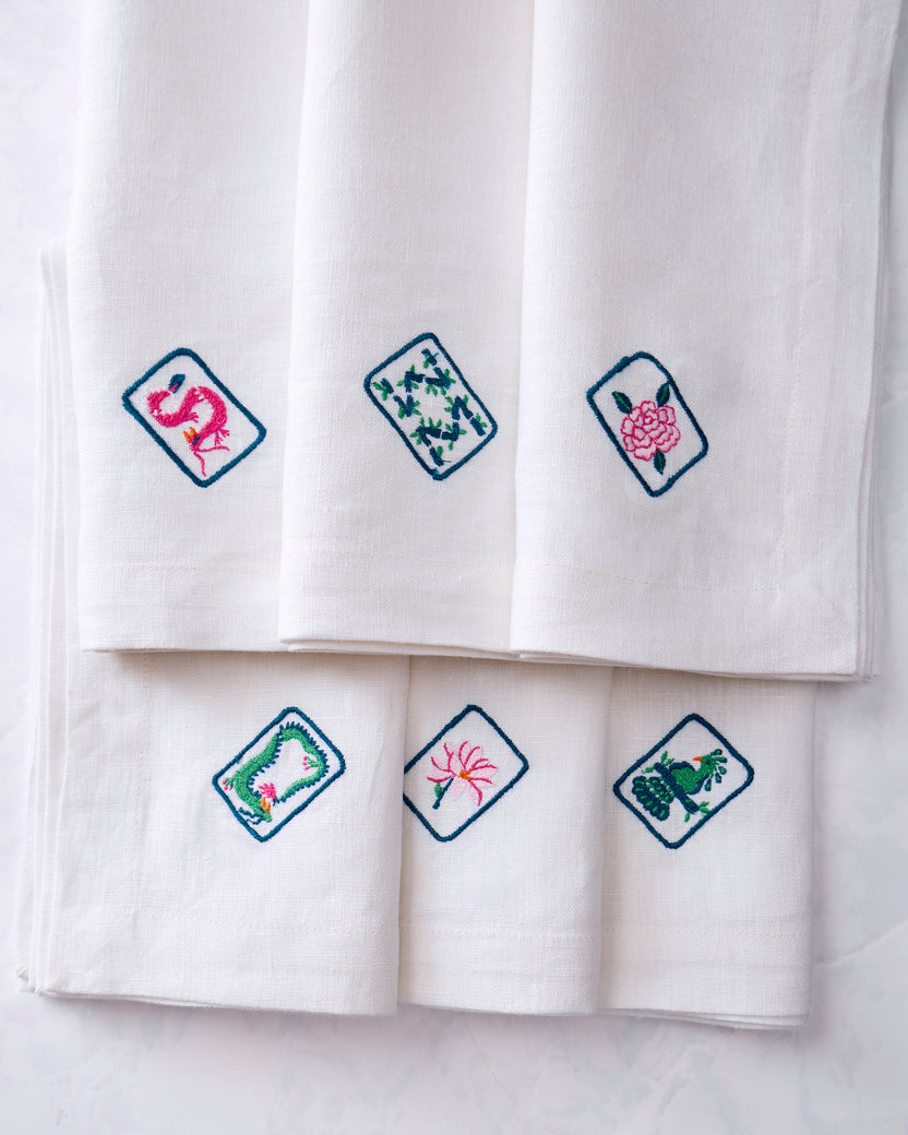 Close-up of six white dinner napkins displaying different colorful embroidered tile motifs, including a pink flamingo, leafy design, pink flower, floral wreath, flower, and green animal motif.