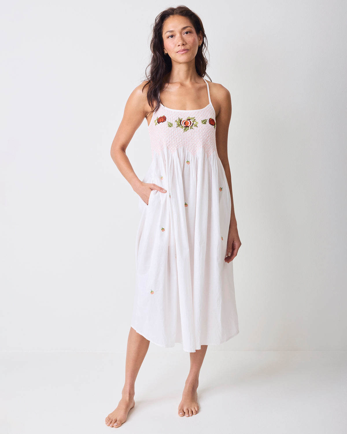 Full view of a white midi nightgown with shoulder straps, an embroidered smocked bodice, scattered pumpkin details, and side pockets.