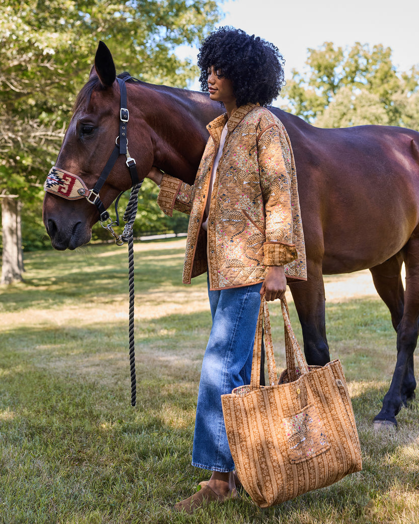 Person carrying a quilted tote bag beside a horse outdoors; the bag has a spotted big-cat print and striped handles.