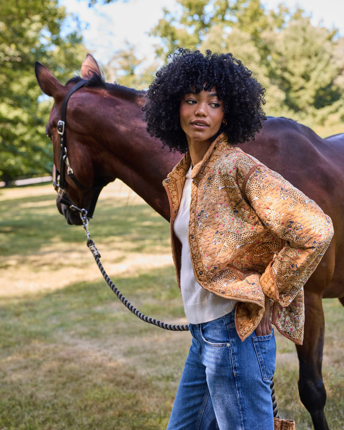 Model wearing the Garden Stripe reversible quilted jacket over a light top with jeans, standing beside a horse.