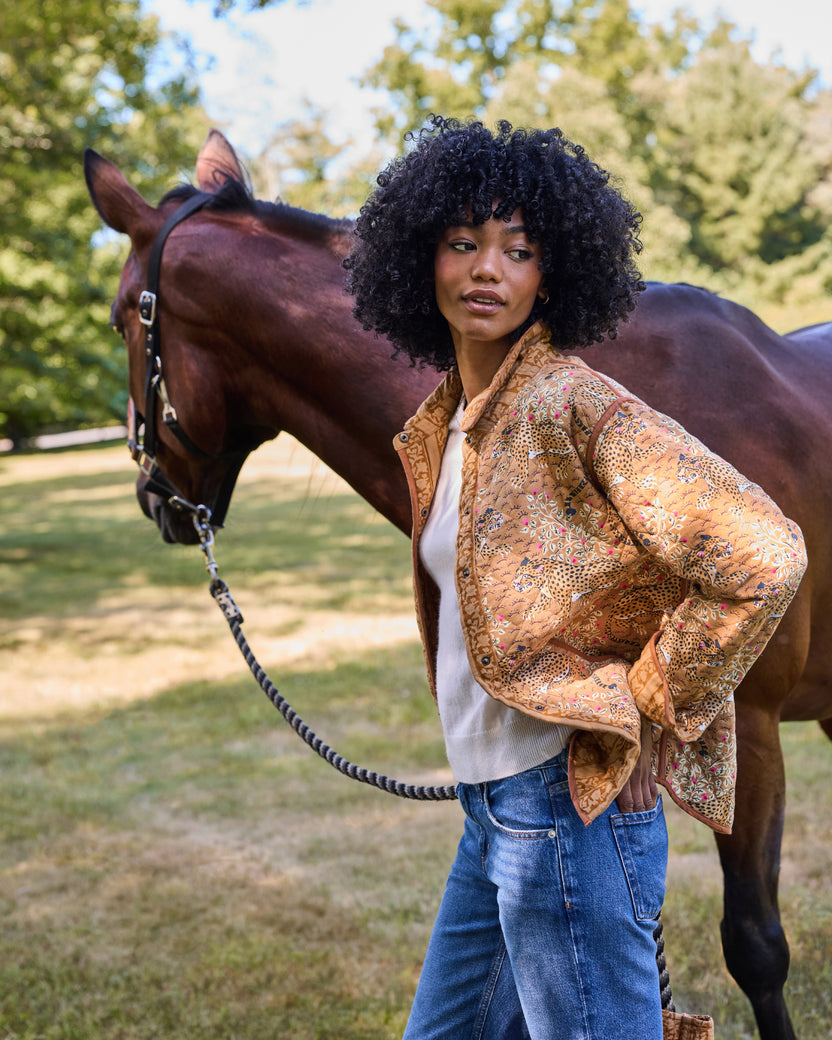 Model wearing the Garden Stripe reversible quilted jacket over a light top with jeans, standing beside a horse.