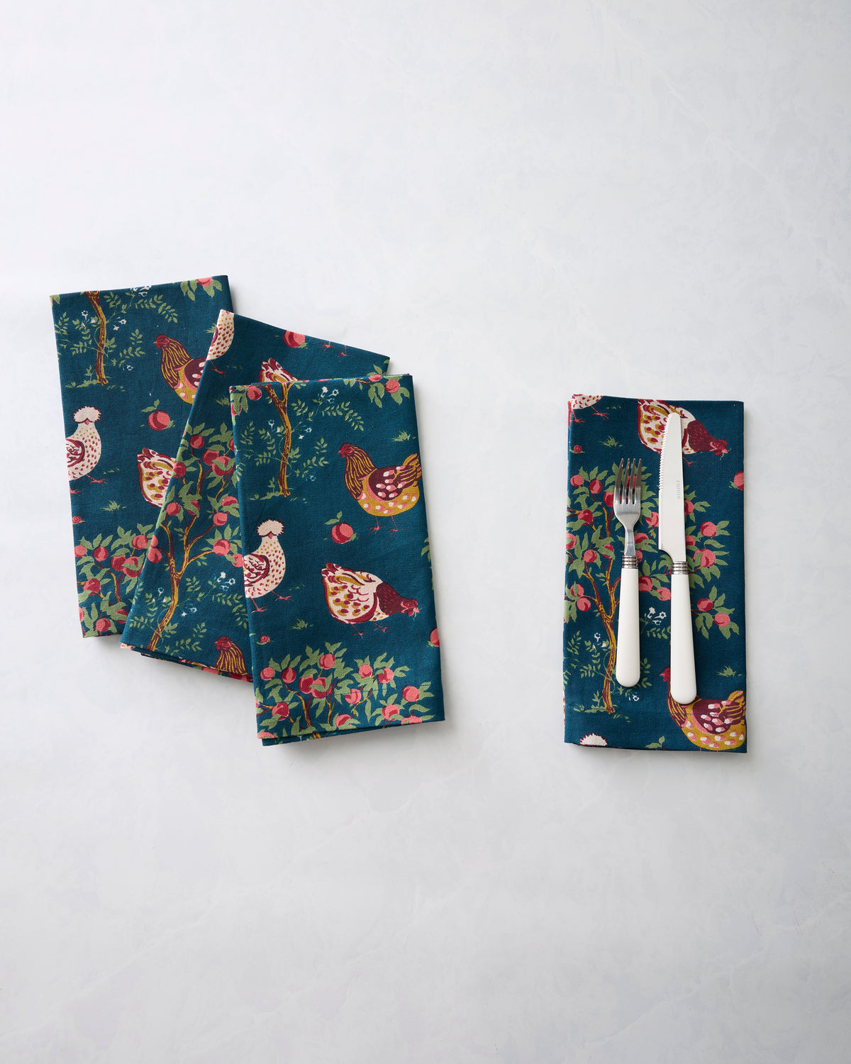 Hen House dinner napkins with chicken-and-apple tree print, including one napkin holding a fork and knife.