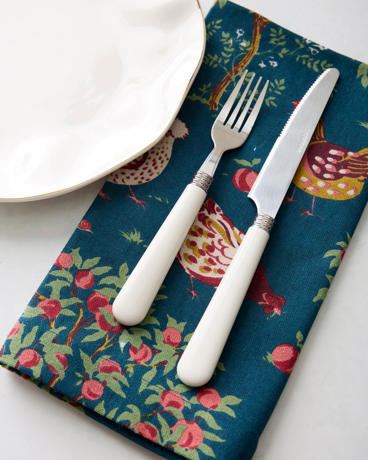 Close-up of a Hen House dinner napkin beside a plate, with a fork and knife resting on the chicken-and-apple print.