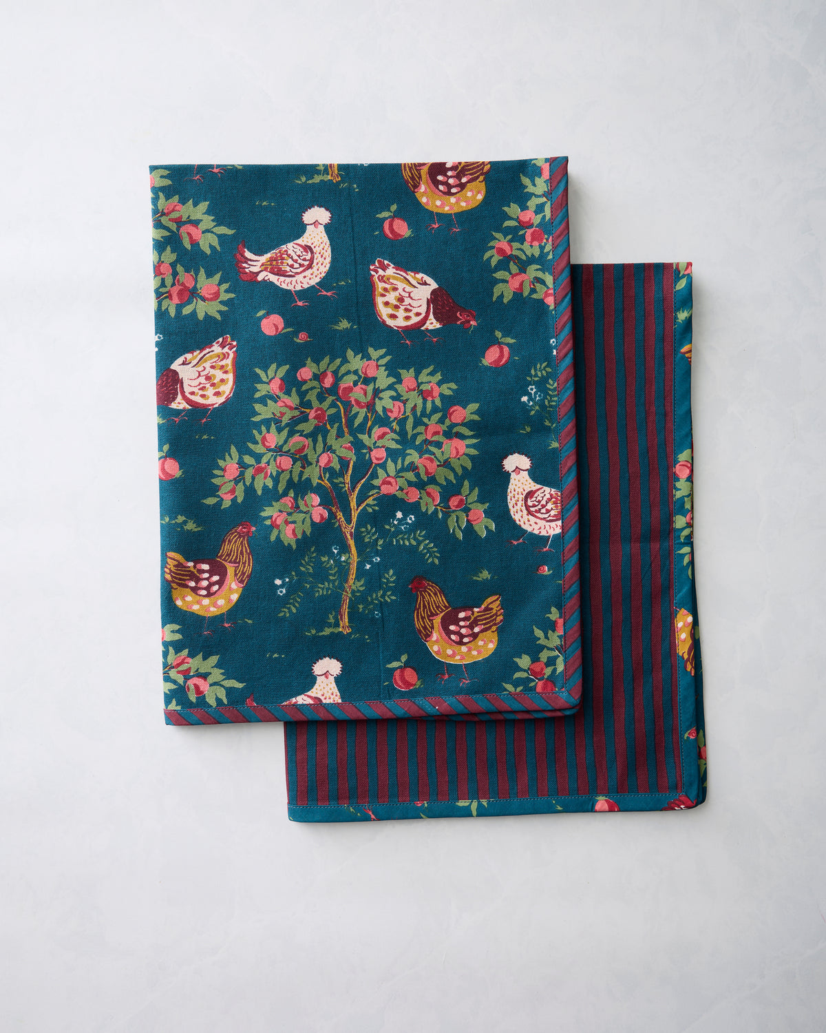 Two overlapping tea towels showing the chicken-and-apple-tree print on one side and the dark teal and burgundy striped reverse on the other.