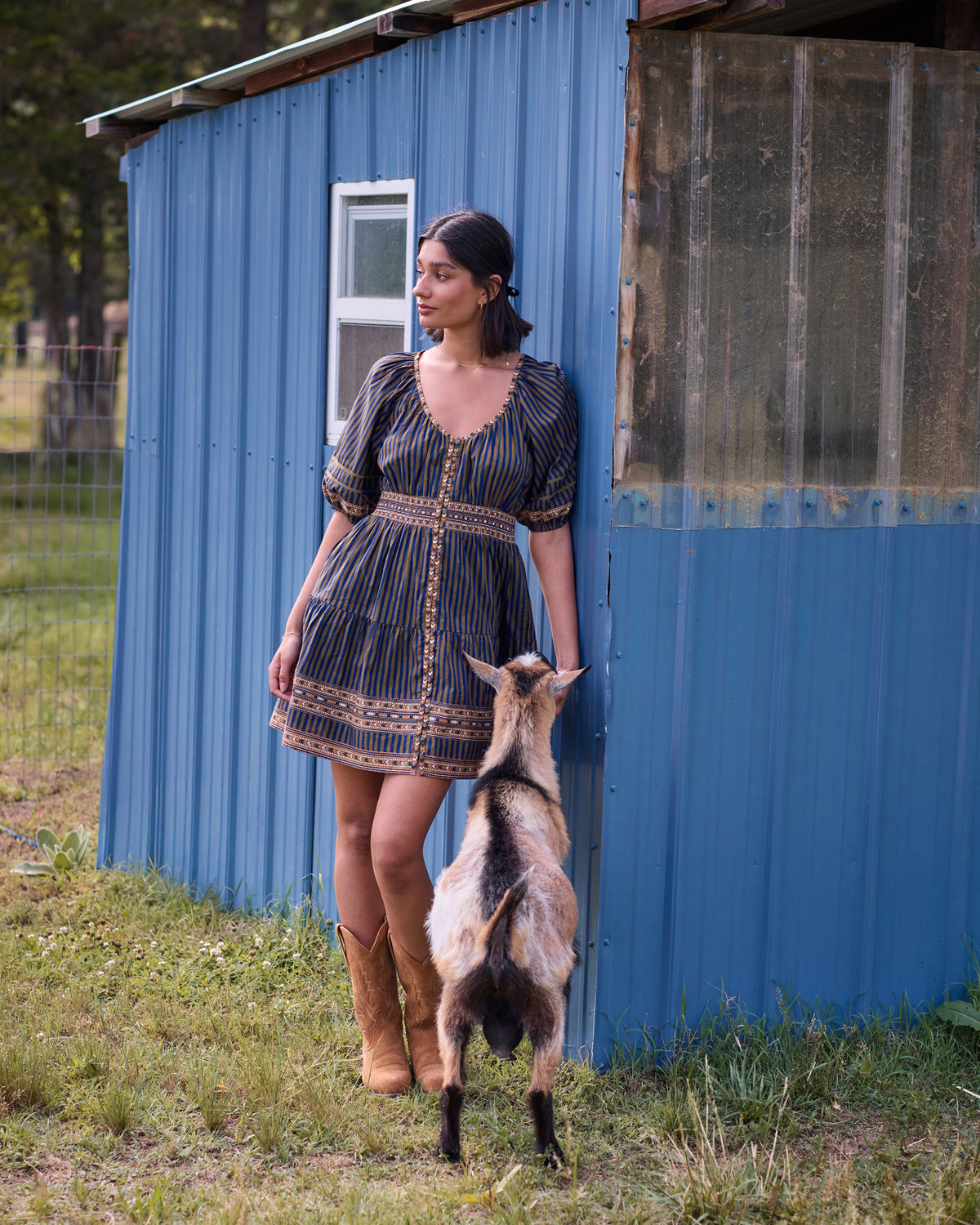 Olive and indigo striped Bryce dress with short puff sleeves, shown beside a blue outbuilding and goat.