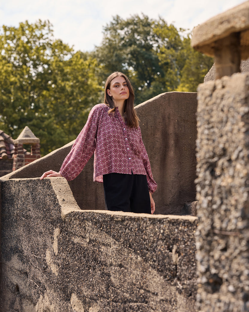 Person wearing a patterned long-sleeve blouse outdoors among stone walls and trees.