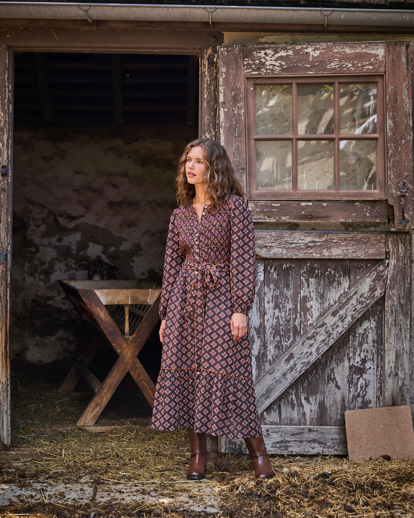 Model wearing the Desert Primrose Shay Dress with a patterned print, long sleeves, waist tie, and ankle boots beside a barn door.